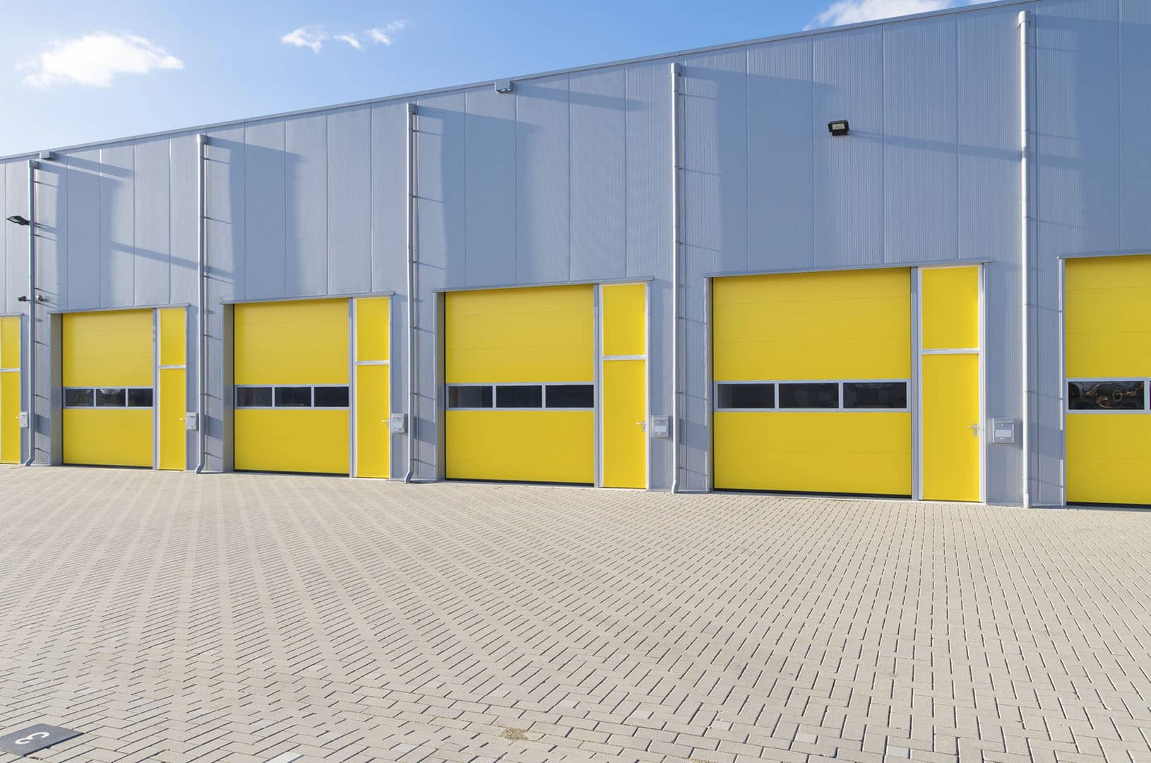 A modern industrial building featuring several large yellow and gray commercial overhead doors and a paved brick courtyard in front, all set beneath a blue sky with scattered clouds.