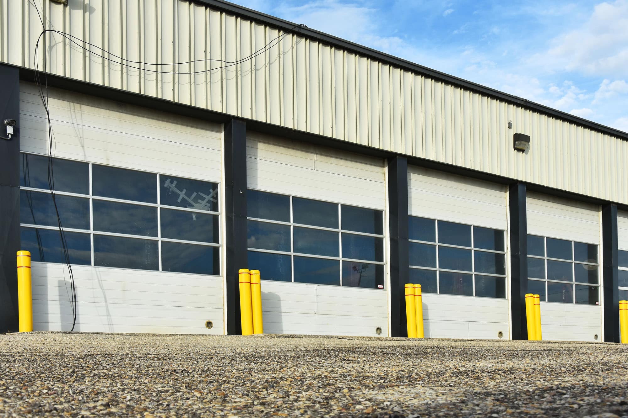 A row of closed white garage doors with glass panels on a commercial building, designed for overhead door energy efficiency, with yellow bollards in front and a cloudy blue sky overhead.