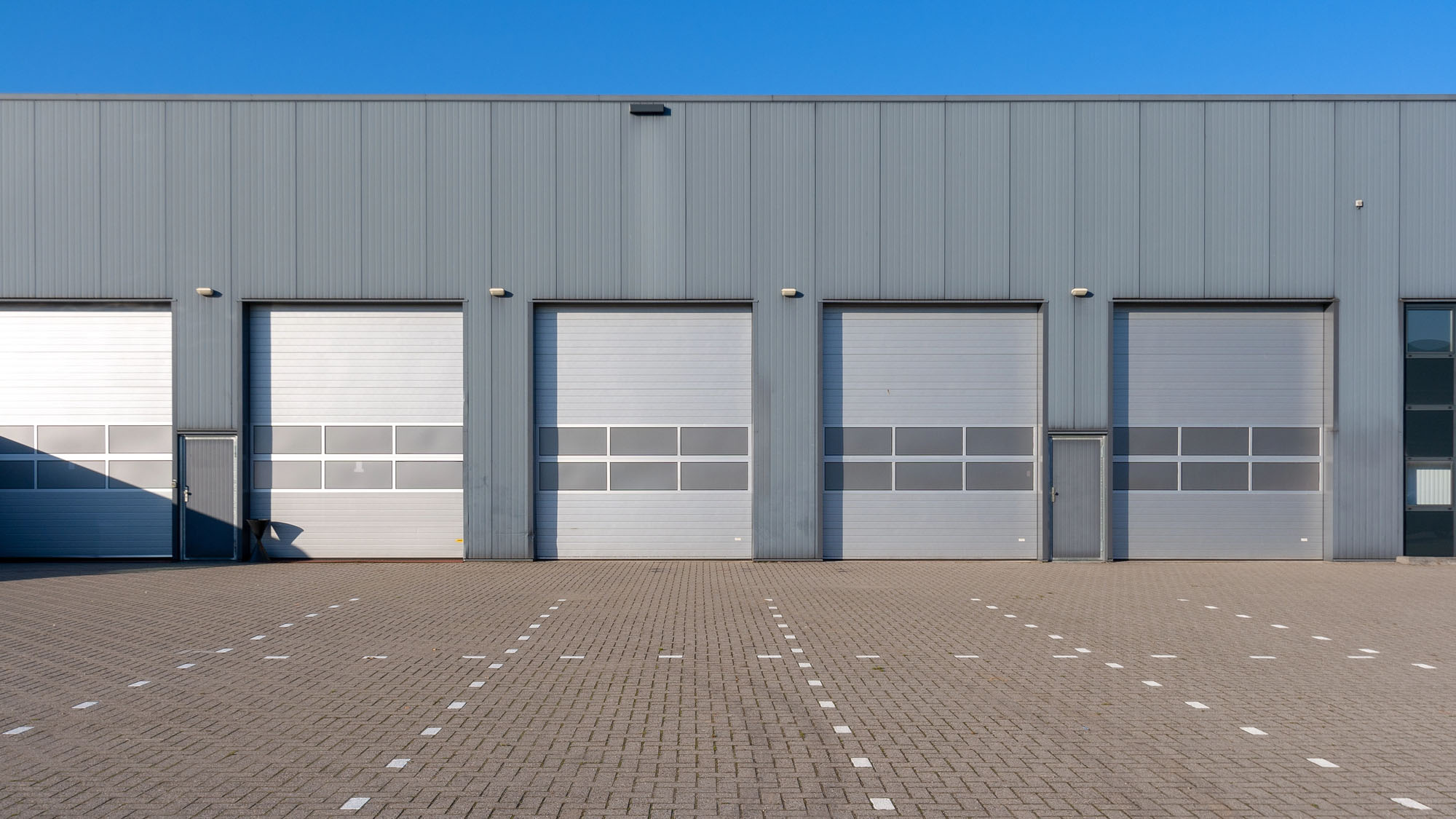 A row of four large, closed High-Speed Overhead Doors with a gray metal exterior and two single doors, set against a clear blue sky and an empty paved lot with lined parking spaces.