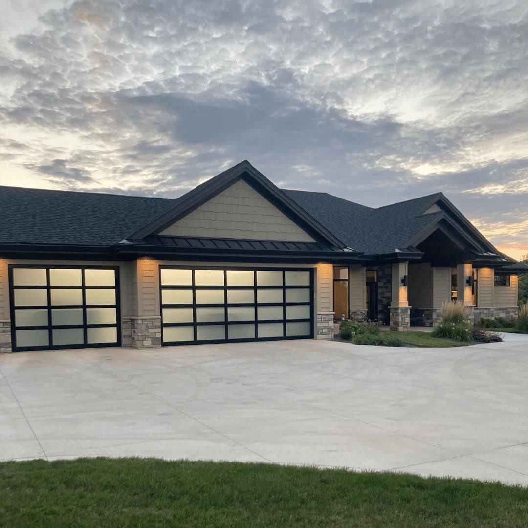 Modern single-story house with three large frosted glass garage doors, stone and wood accents, a wide driveway, and outdoor lights on at dusk under a cloudy sky.