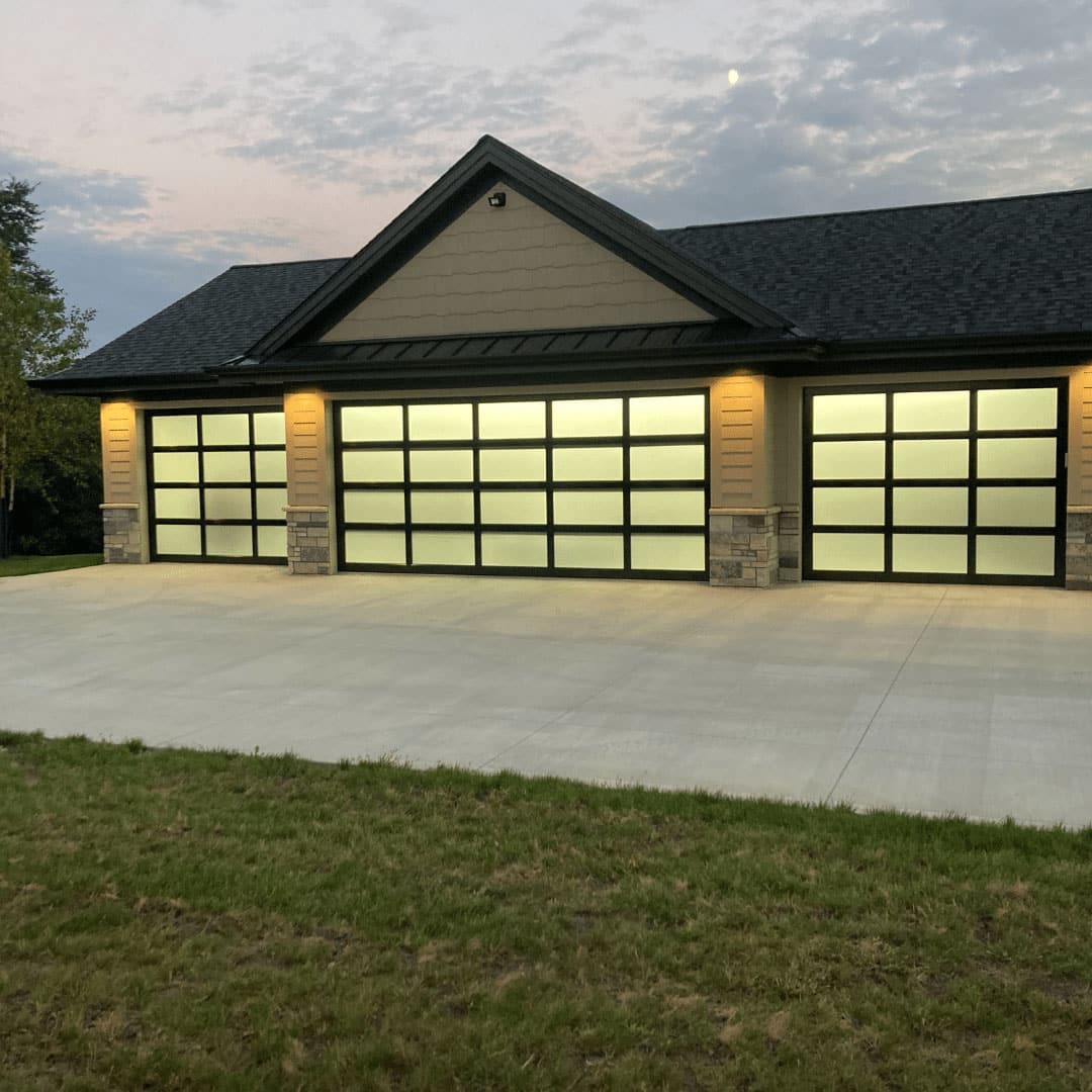 quality-overhead-door-44 Modern house with three large, frosted residential garage doors, exterior lights on, and a wide concrete driveway in front. The sky is cloudy with some sunset light, and there is grass in the foreground.