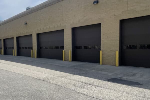 A row of large, closed, dark garage doors with yellow bollards in front, set in a beige brick building under a cloudy sky. The ground is paved with asphalt and concrete.