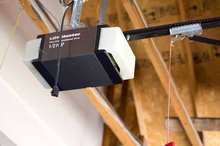 A garage door opener is mounted on the ceiling with exposed wooden beams. A black power cord hangs down, and a red emergency release cord is visible.