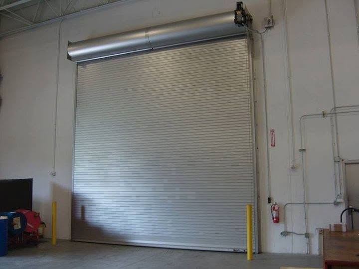A large, silver industrial roll-up garage door is closed inside a warehouse. The walls are white, with visible wiring, pipes, and a fire extinguisher mounted nearby. Two yellow posts stand in front of the door.