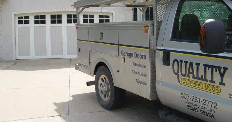 A service truck labeled QUALITY Overhead Door is parked in a driveway near a white residential garage door. The truck advertises garage door services for residential, commercial, and opener installations.