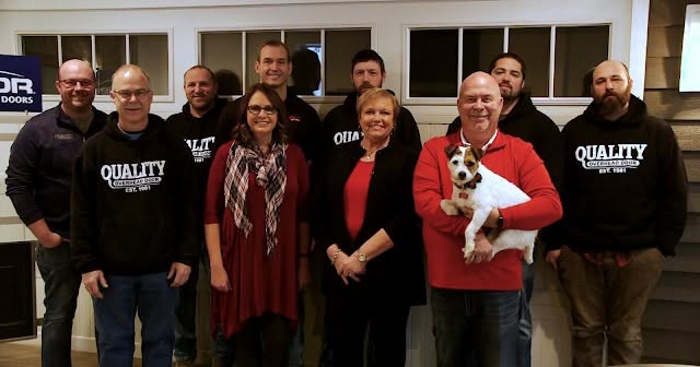 A group of ten adults, including two women, pose indoors. One man in front wears red and holds a small dog. Several men wear black Quality Construction hoodies. They smile, standing in front of windows.
