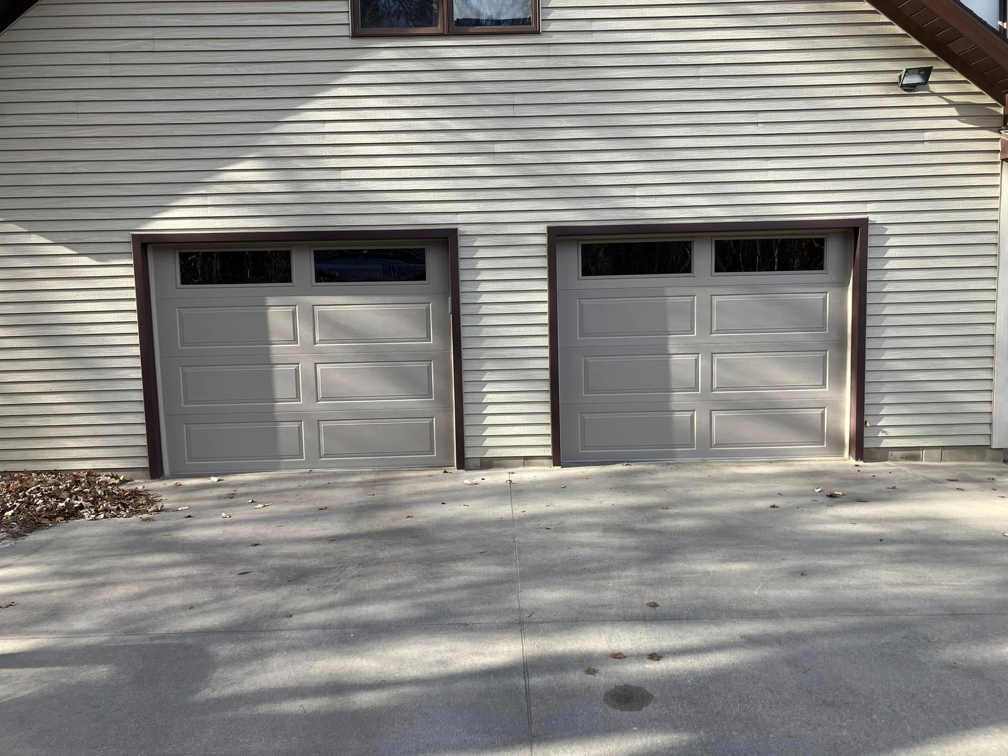 Two closed beige garage doors with small rectangular windows at the top, set in a light-colored siding house. Shadows and leaves are visible on the concrete driveway in front.