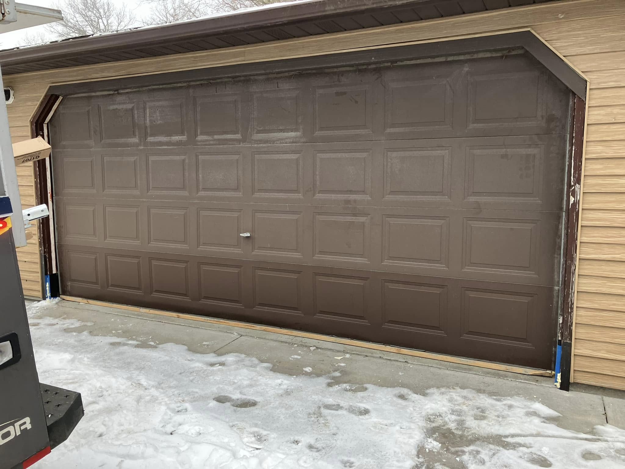 A brown garage door is off its tracks and leaning forward, with snow and ice on the ground in front of it. The door frame and siding are tan-colored.