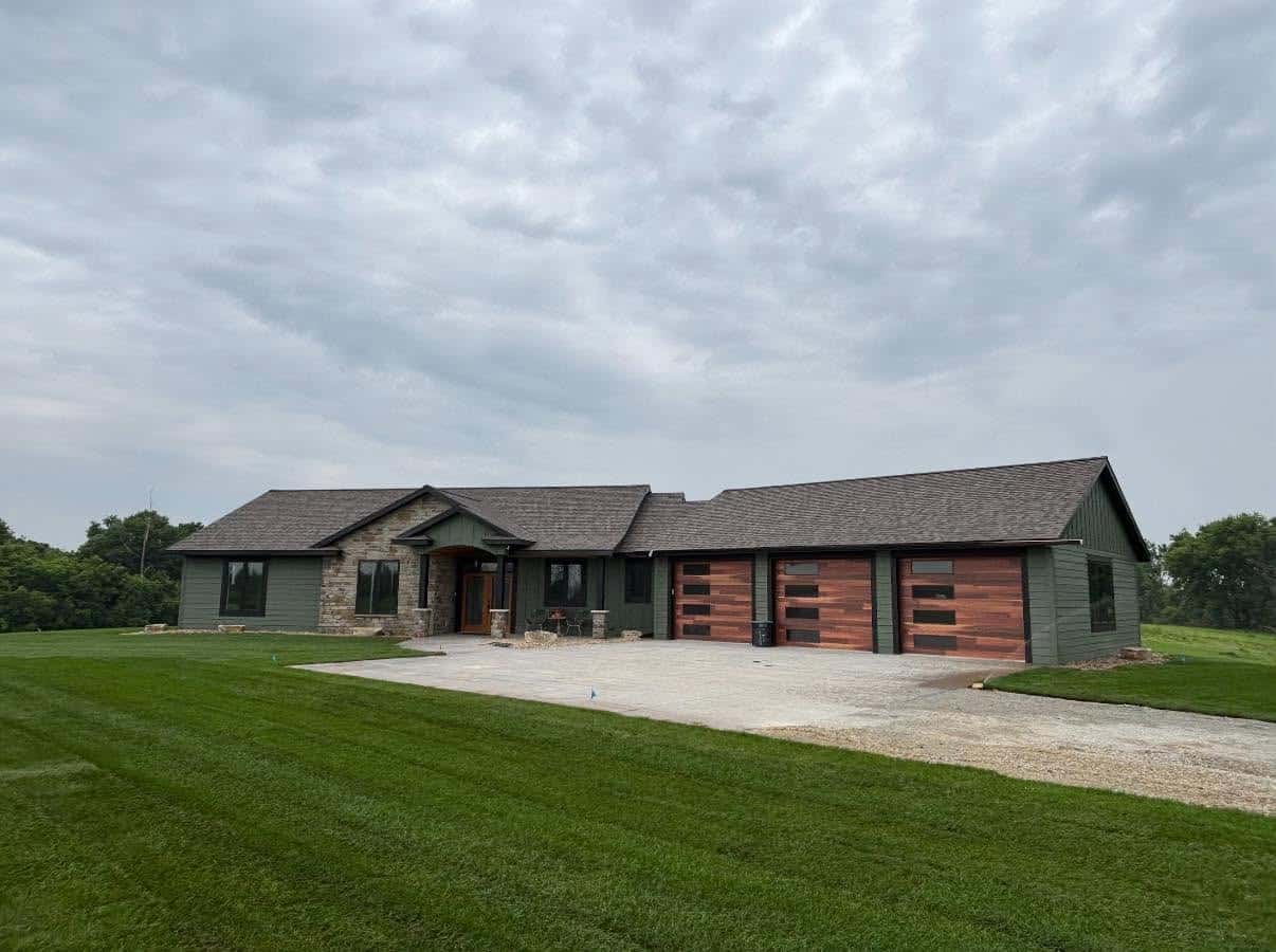 Single-story green house with stone accents, three-car garage with wood panel doors, set on a large, neatly mowed lawn under a cloudy sky.
