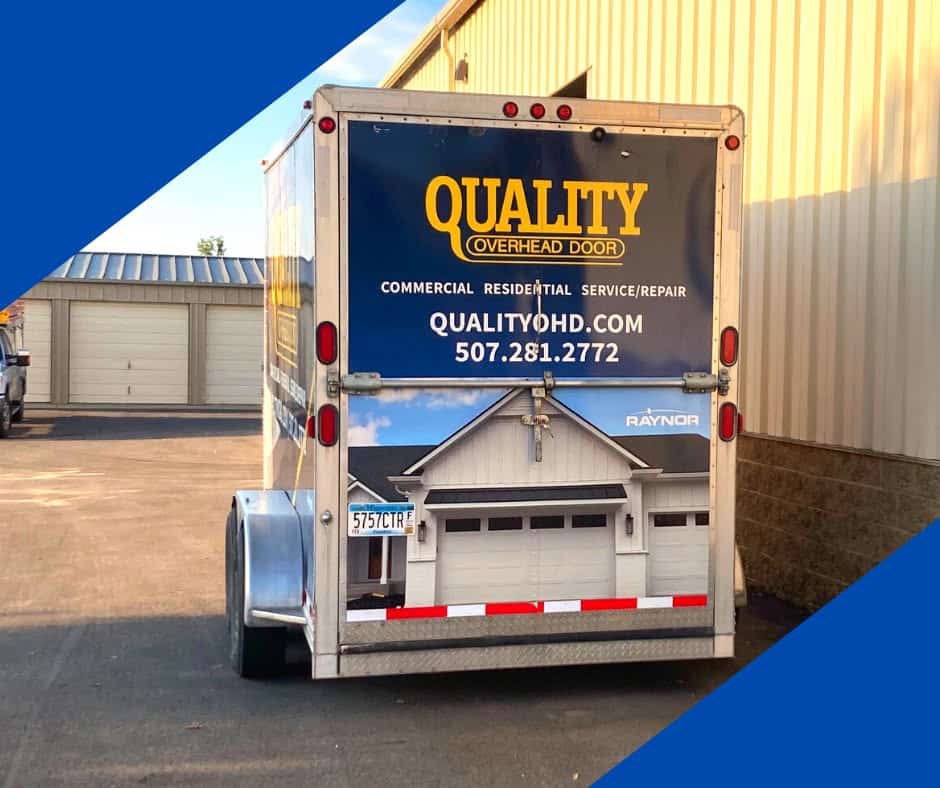 A trailer with a Quality Overhead Door ad showing a garage door, contact info, and phone number is parked next to a beige building.