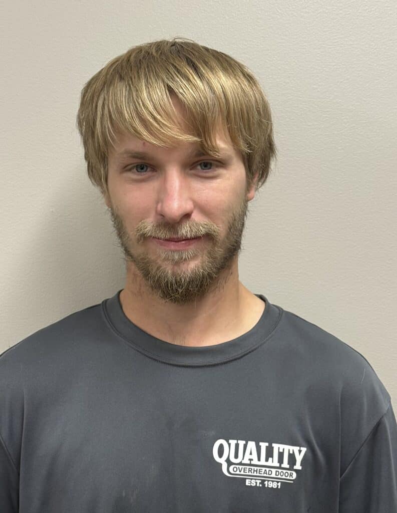 A man with blond hair and a beard wearing a gray shirt that says QUALITY OVERHEAD DOOR EST. 1981, standing in front of a plain light-colored wall.