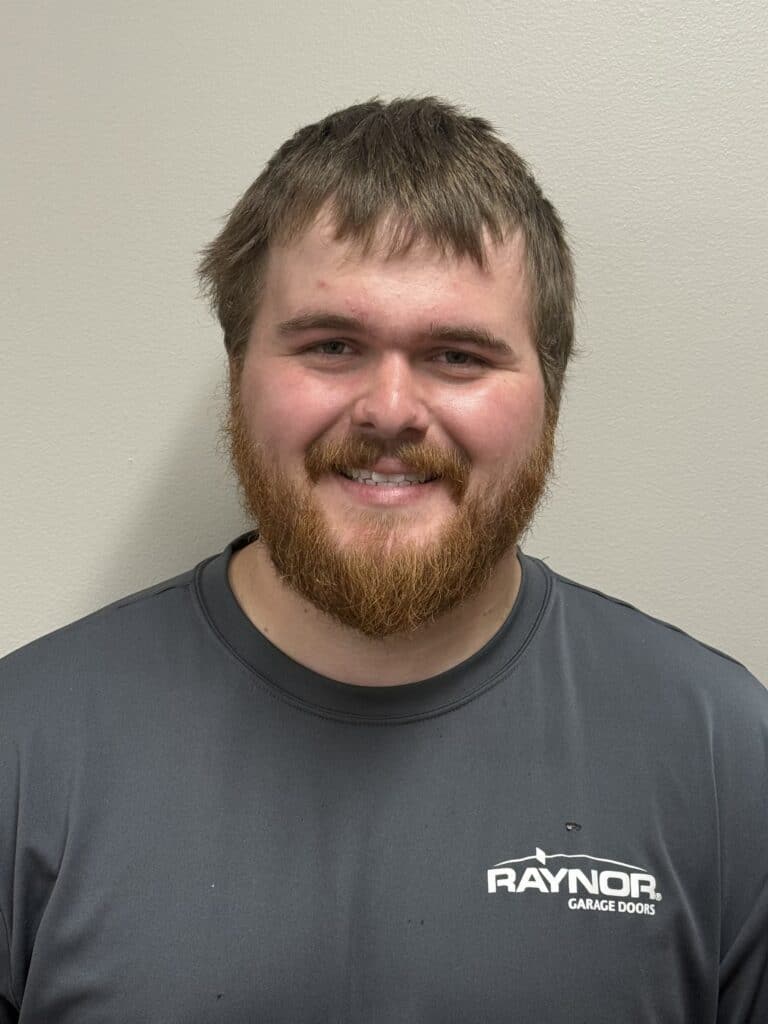 A man with a short brown beard and hair smiles at the camera. He is wearing a gray Raynor Garage Doors shirt and stands against a plain light-colored wall.