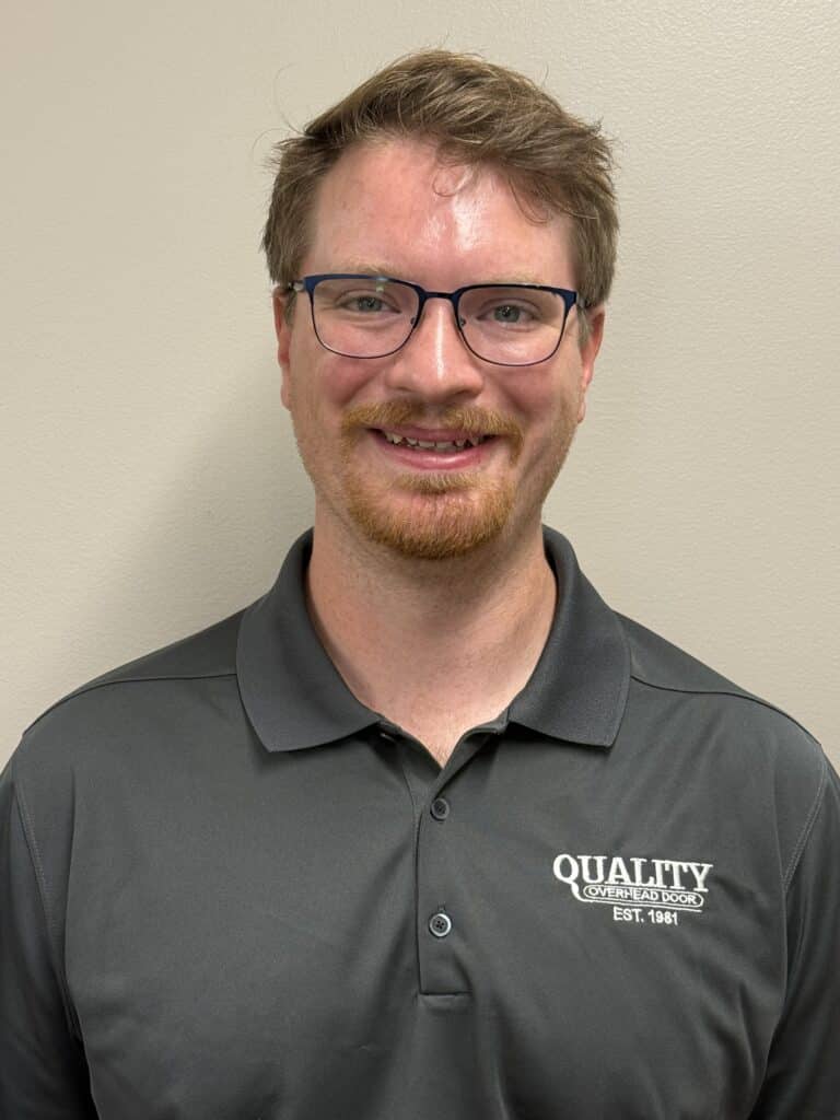 A man with short light brown hair, glasses, and a trimmed beard smiles at the camera. He is wearing a dark gray polo shirt with the words QUALITY (OVERHEAD DOORS) EST. 1981 embroidered on it.
