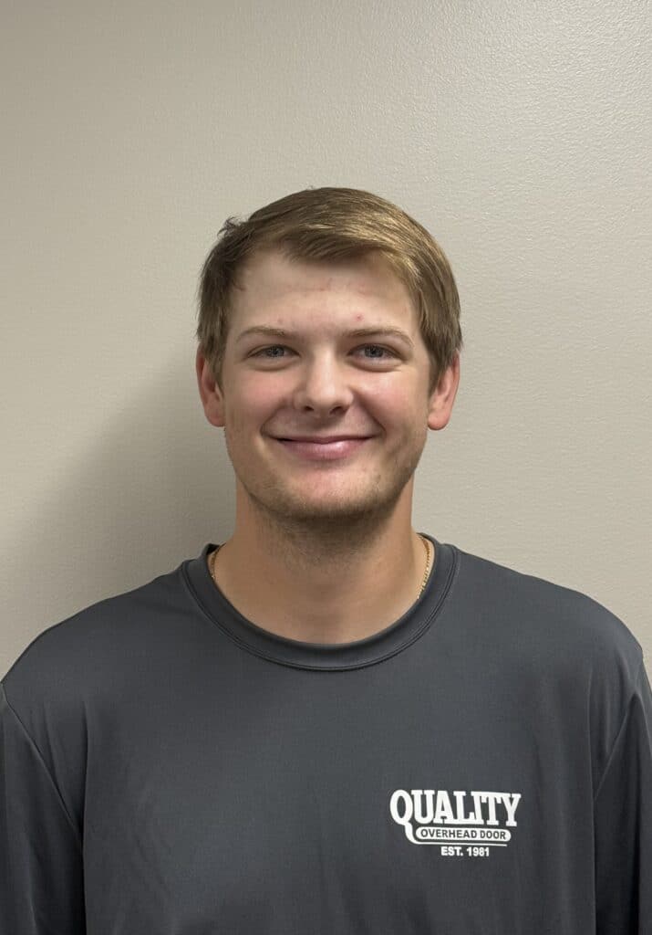 A young man with short light brown hair smiles at the camera. He wears a gray Quality Overhead Door shirt and stands against a plain beige wall.