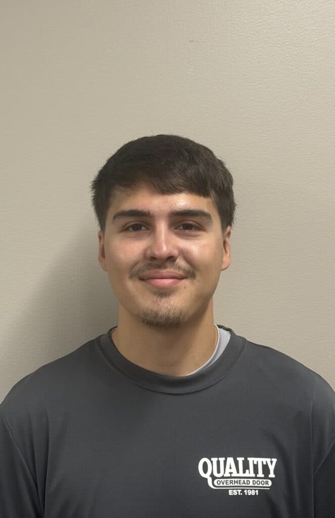 A young man with short dark hair and a trimmed beard smiles at the camera. He is wearing a dark gray Quality Overhead Door shirt and stands against a plain beige wall.
